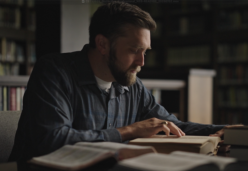 Bearded man in a dark shirt reading a book at a table surrounded by open books in a dimly lit library.
