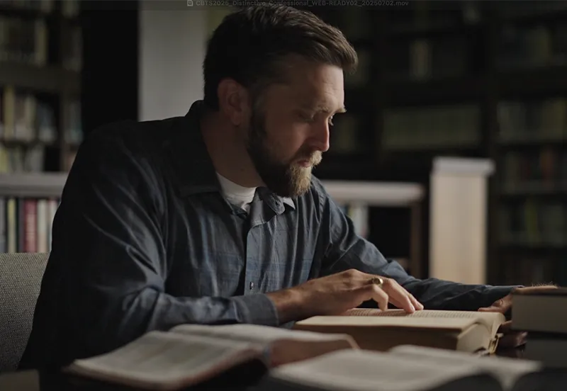 Bearded man in a dark shirt reading a book at a table surrounded by open books in a dimly lit library.