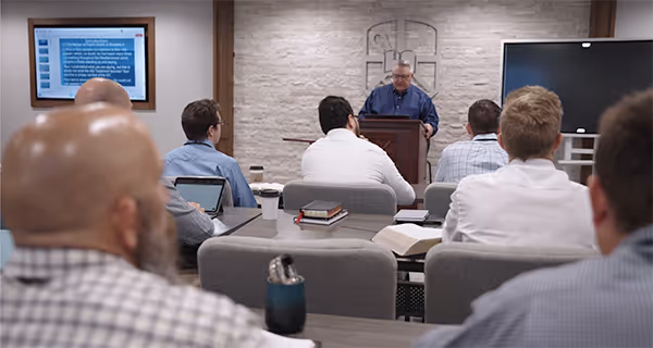 Man reading at a podium in a classroom setting with an audience seated facing him, and a presentation slide displayed on a screen.