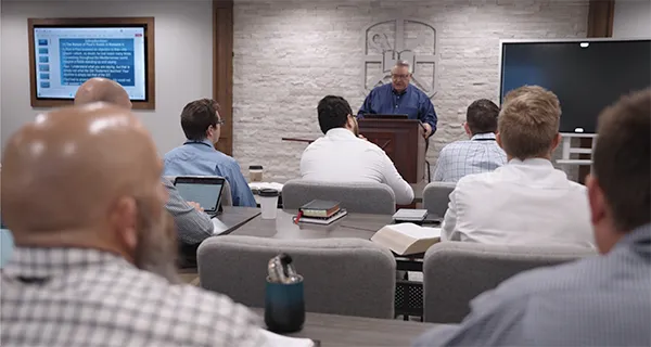 Man reading at a podium in a classroom setting with an audience seated facing him, and a presentation slide displayed on a screen.