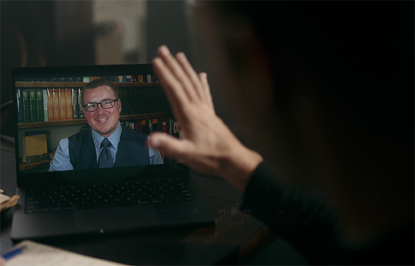 Person waving at a man in glasses and a vest who is smiling on a laptop screen during a video call in a dimly lit room.