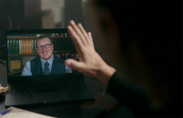 Person waving at a man in glasses and a vest who is smiling on a laptop screen during a video call in a dimly lit room.