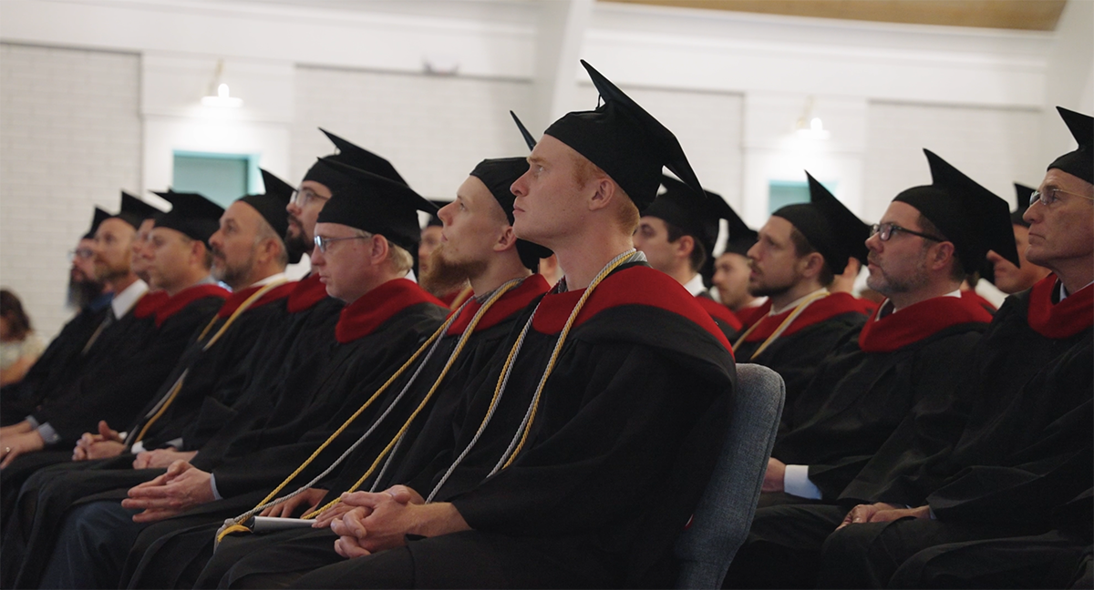 Row of male graduates in black gowns and caps with red stoles attentively watching a ceremony indoors.