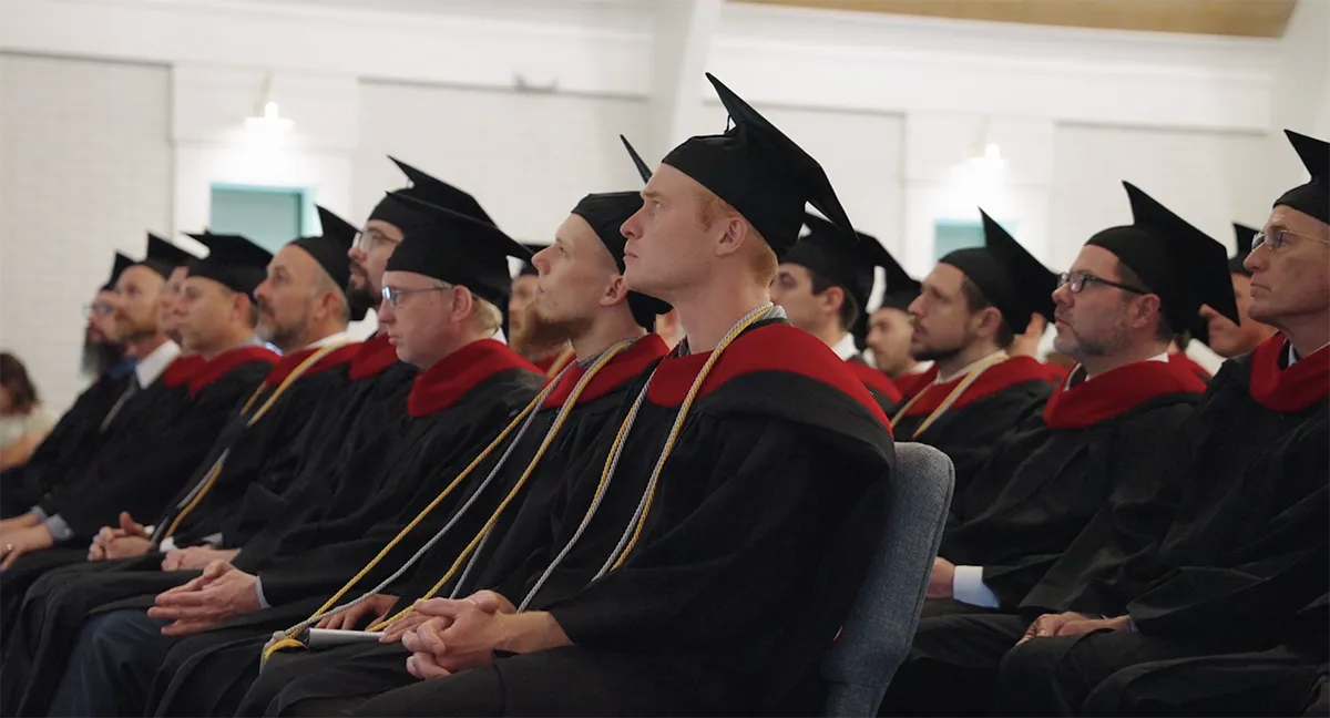Row of male graduates in black gowns and caps with red stoles attentively watching a ceremony indoors.