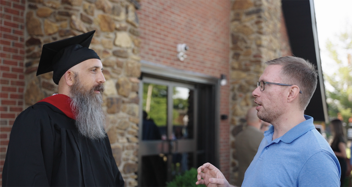 Bearded man in graduation cap and gown talking to a man in a blue polo shirt outside a building with stone and brick walls.