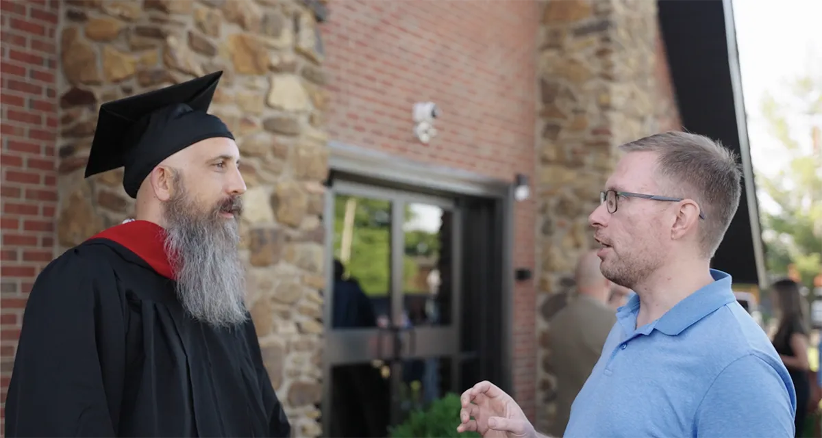 Bearded man in graduation cap and gown talking to a man in a blue polo shirt outside a building with stone and brick walls.