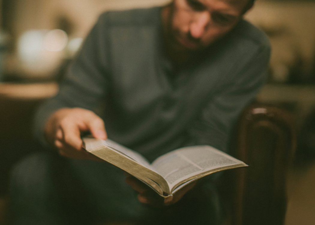 Man sitting and reading a thick book with blurred background.