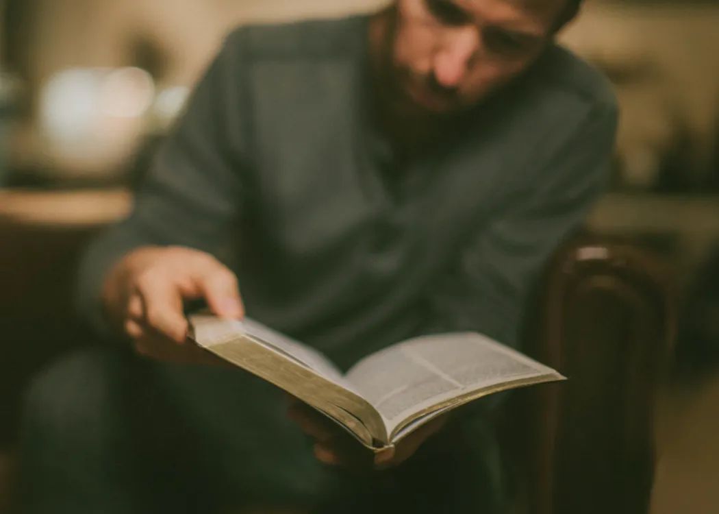 Man sitting and reading a thick book with blurred background.
