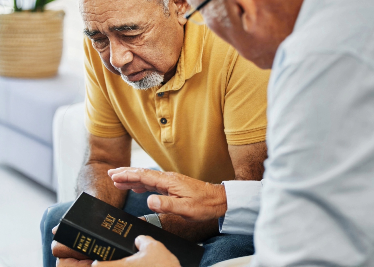 Two elderly men sitting closely, one holding a black Holy Bible while the other gestures with his hand.