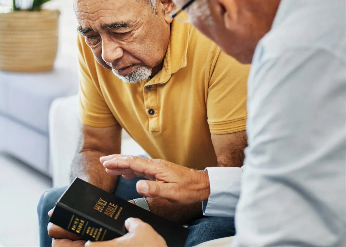Two elderly men sitting closely, one holding a black Holy Bible while the other gestures with his hand.