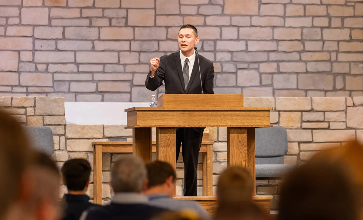 Man in a suit speaking at a wooden podium in front of an audience inside a stone-walled room.
