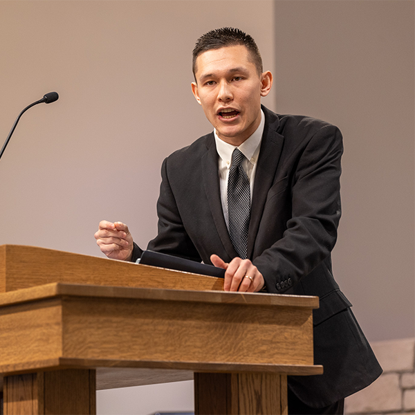 Man in a black suit speaking passionately behind a wooden podium with a microphone.