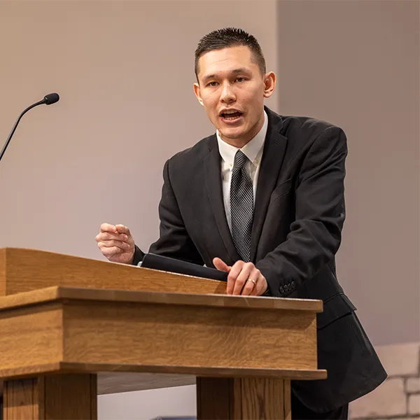 Man in a black suit speaking passionately behind a wooden podium with a microphone.