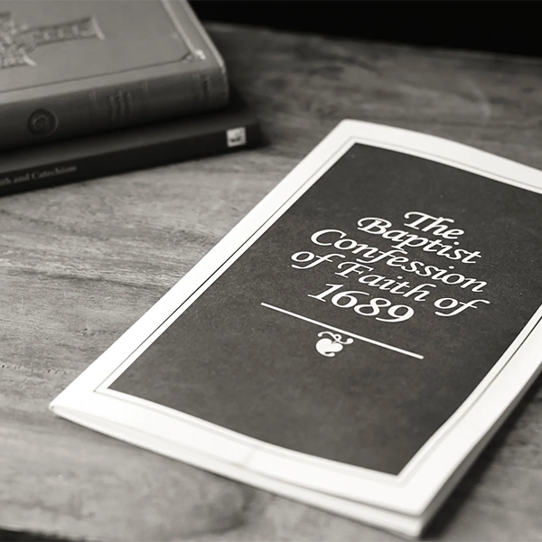 A black and white booklet titled 'The Baptist Confession of Faith of 1689' lying on a wooden surface with two closed books in the background.