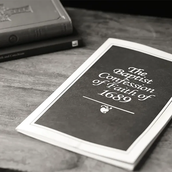 A black and white booklet titled 'The Baptist Confession of Faith of 1689' lying on a wooden surface with two closed books in the background.