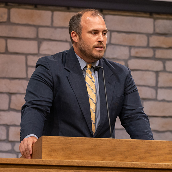 Man in a dark suit with a yellow tie speaking at a wooden podium with a microphone, against a stone wall background.