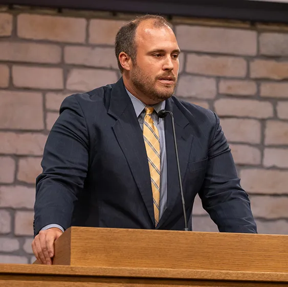 Man in a dark suit with a yellow tie speaking at a wooden podium with a microphone, against a stone wall background.