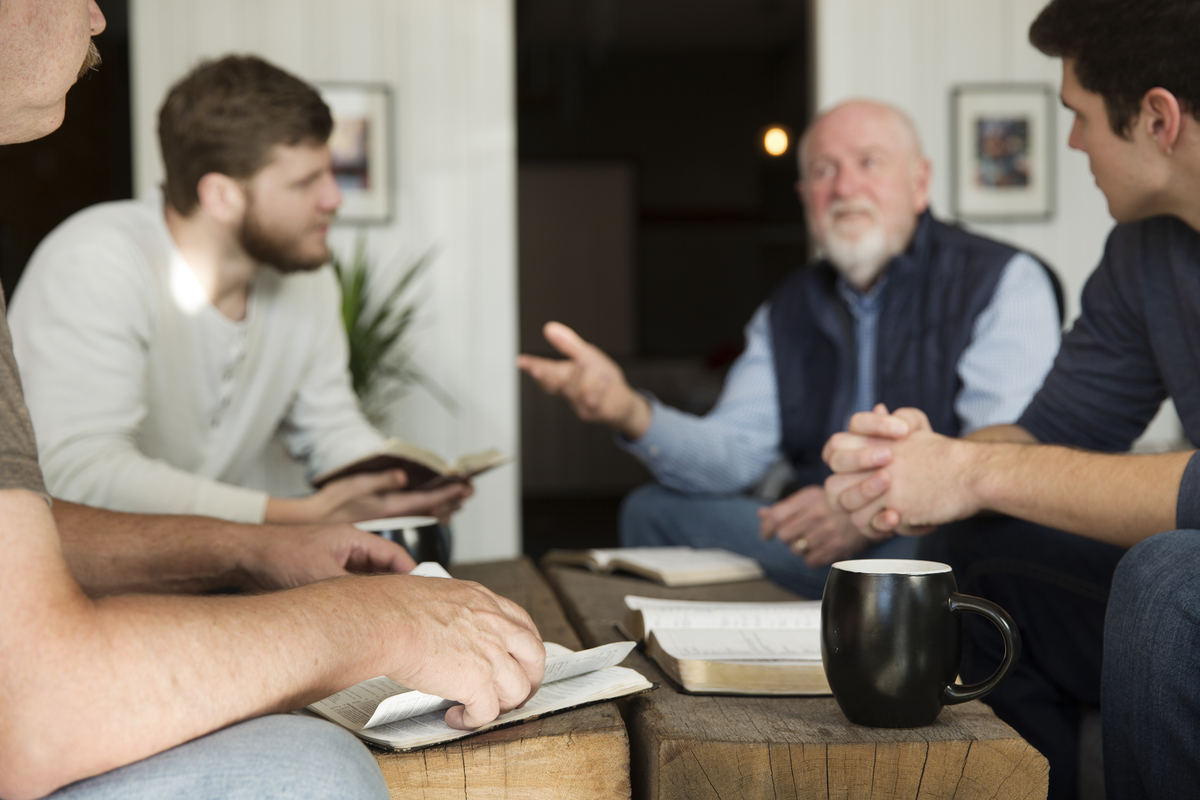 Group of men sitting around a wooden table with open books and a black coffee mug, engaged in conversation.