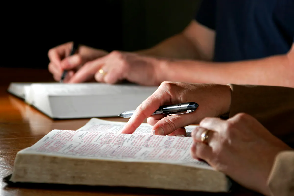 Two people reading and writing in open Bibles on a wooden table.