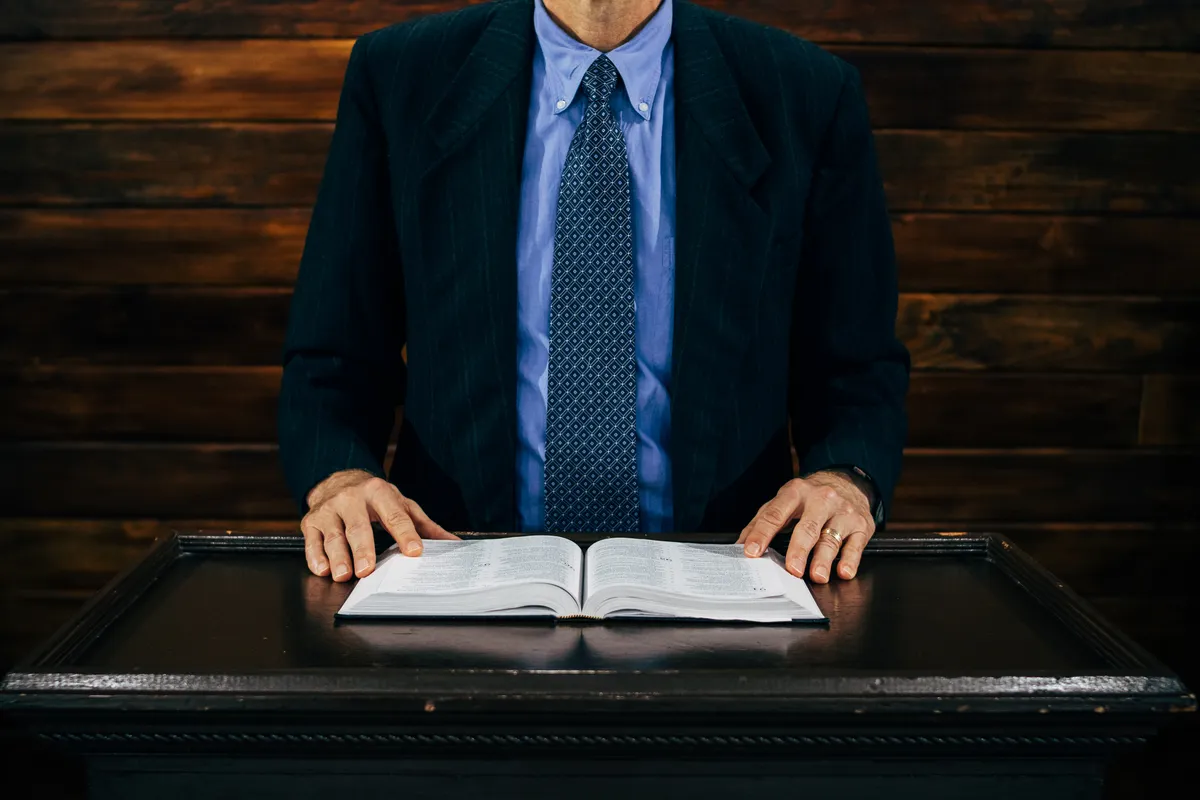 Man in a suit and tie standing behind a lectern with an open book.