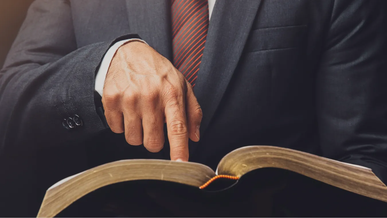 Person in a suit and red striped tie pointing at a page in an open book.