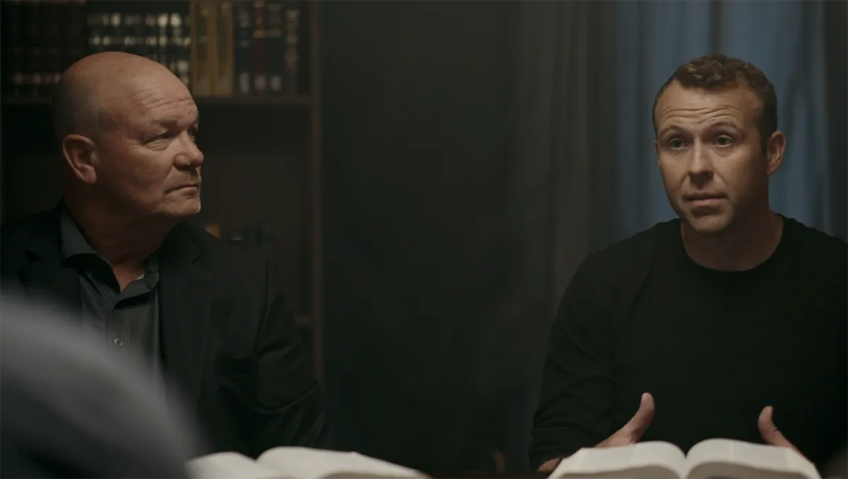 Two men sitting at a table with open books, one older man in a dark suit looking to the side and a younger man in a black shirt speaking.
