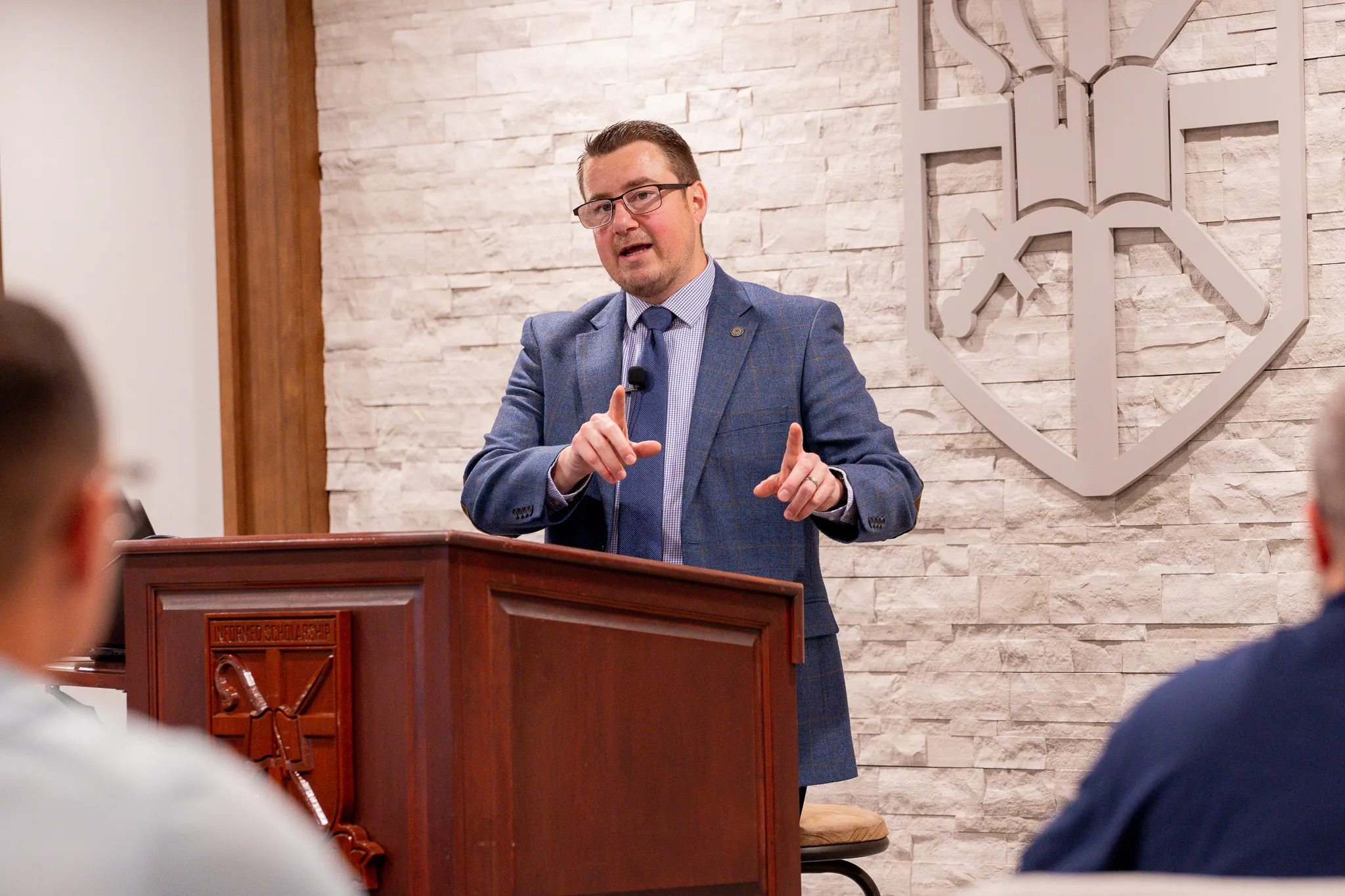 Man in glasses and blue suit speaking behind a wooden podium with a shield emblem on a stone wall background.