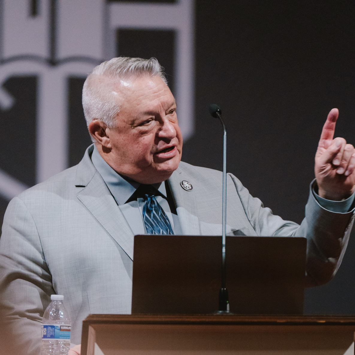 Man in a light gray suit speaking at a podium with a microphone, gesturing with his right hand raised.