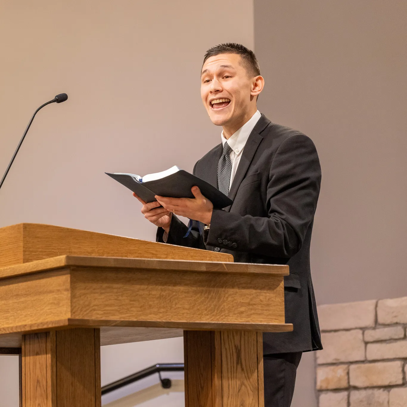 Man in a black suit speaking enthusiastically while holding an open book at a wooden podium with a microphone.