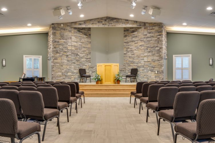 Interior of a church sanctuary with rows of brown chairs facing a raised podium flanked by two chairs and stone wall backdrop.