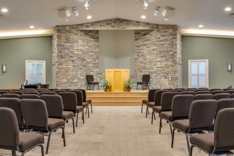 Interior of a church sanctuary with rows of brown chairs facing a raised podium flanked by two chairs and stone wall backdrop.
