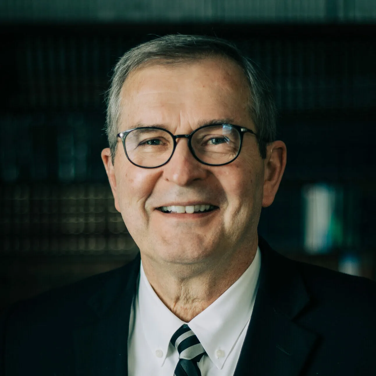 Smiling middle-aged man wearing glasses, a white shirt, striped tie, and black suit jacket, with a blurred dark background.