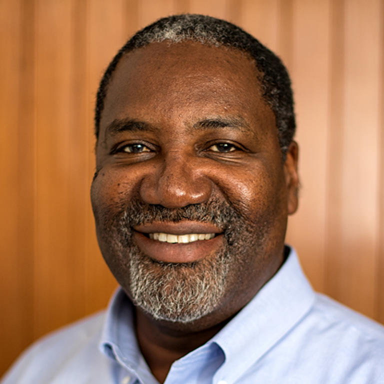 Smiling middle-aged man with salt-and-pepper hair and beard, wearing a light blue collared shirt against a wooden background.