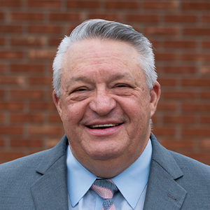 Smiling older man with gray hair wearing a gray suit, blue shirt, and striped tie in front of a brick wall.