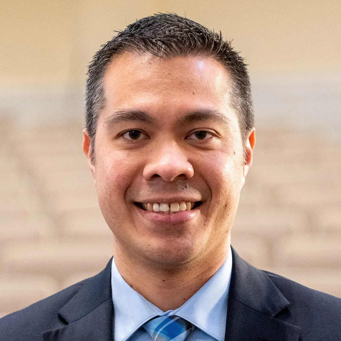 Smiling man with short black hair wearing a blue shirt, plaid tie, and dark suit jacket.