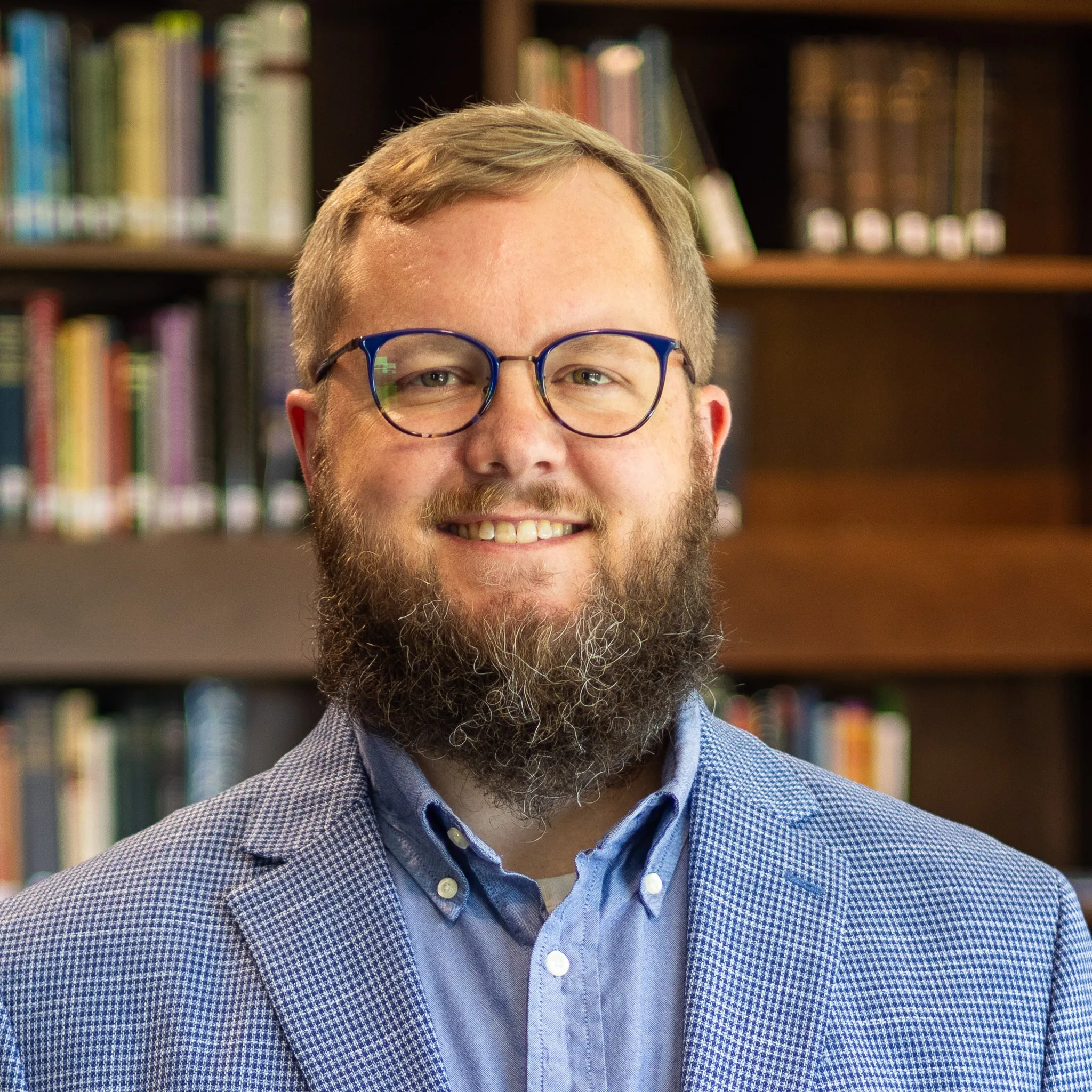 Smiling man with glasses and a full beard wearing a blue checkered blazer and shirt, standing in front of a bookshelf.