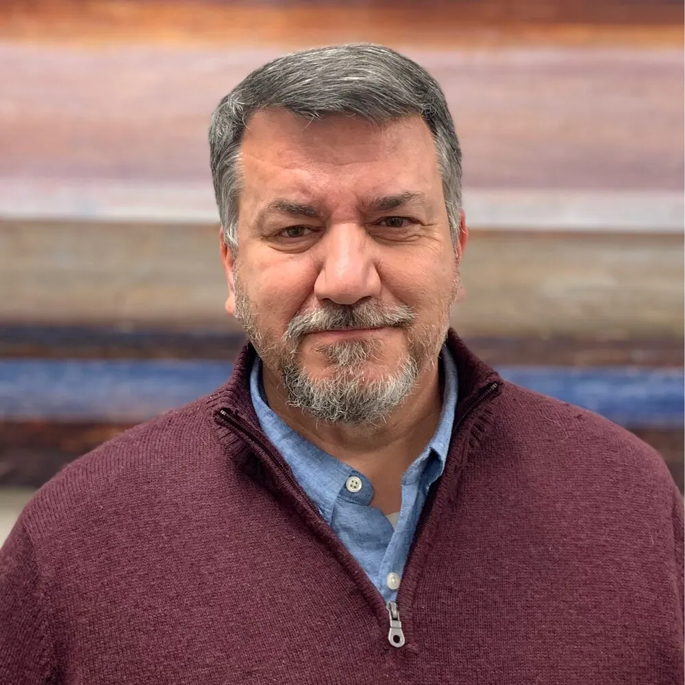 Middle-aged man with gray hair and beard wearing a maroon zip-up sweater over a blue button-up shirt, standing against a blurred background.