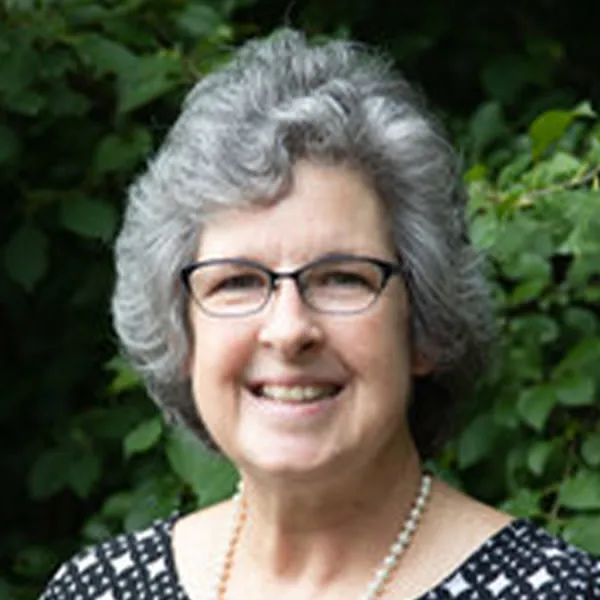 Smiling older woman with curly gray hair, glasses, and a pearl necklace, standing outdoors with green foliage background.