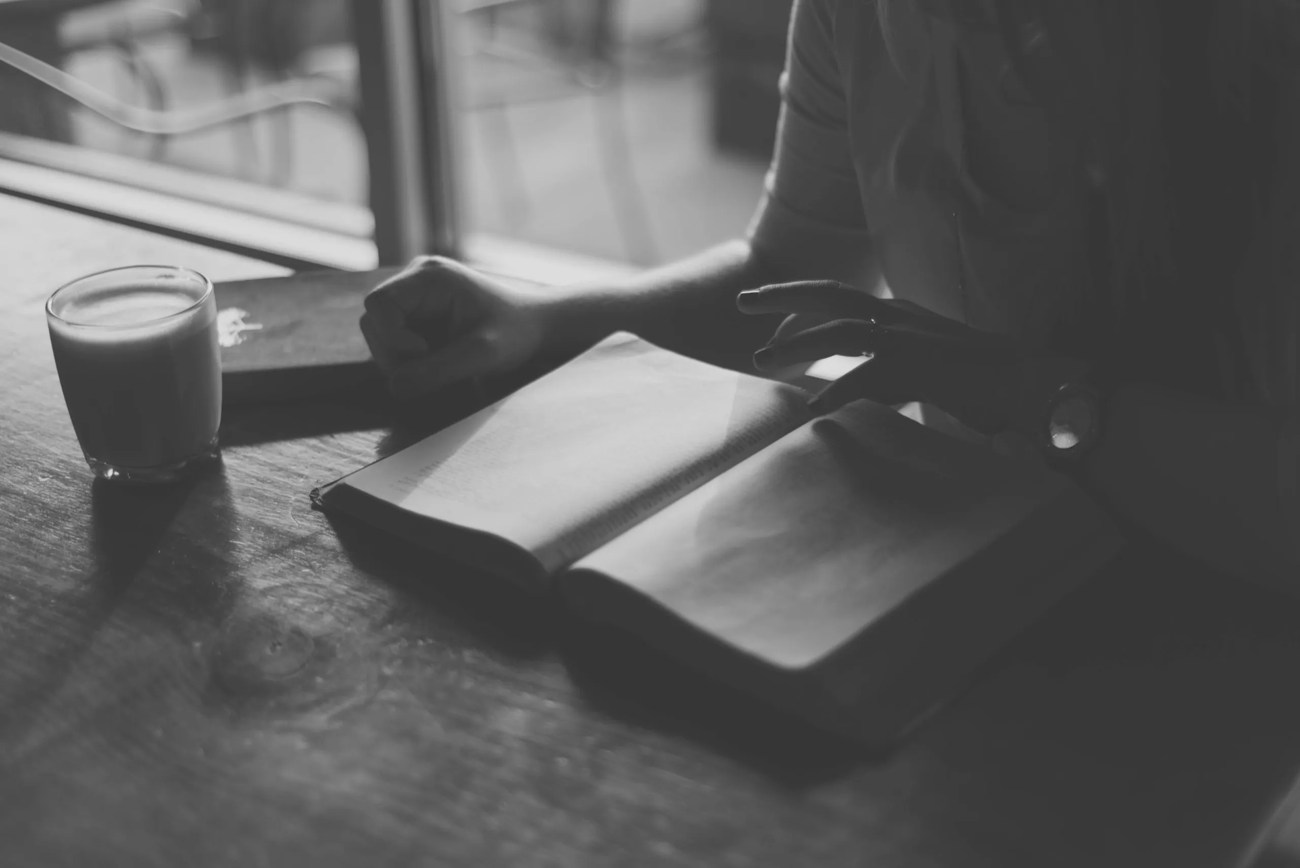 Person reading an open book on a wooden table next to a glass of frothy drink in a softly lit room.