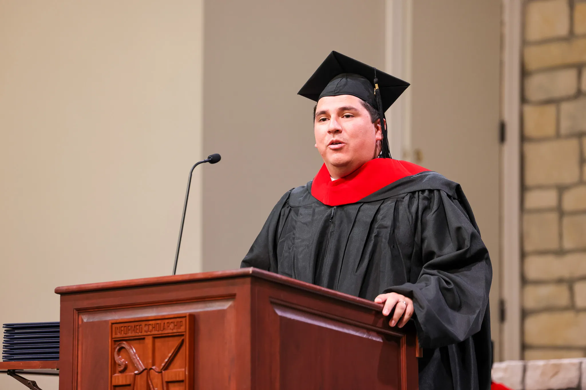 Male graduate in black cap and gown with red hood speaking at a wooden podium with a microphone.
