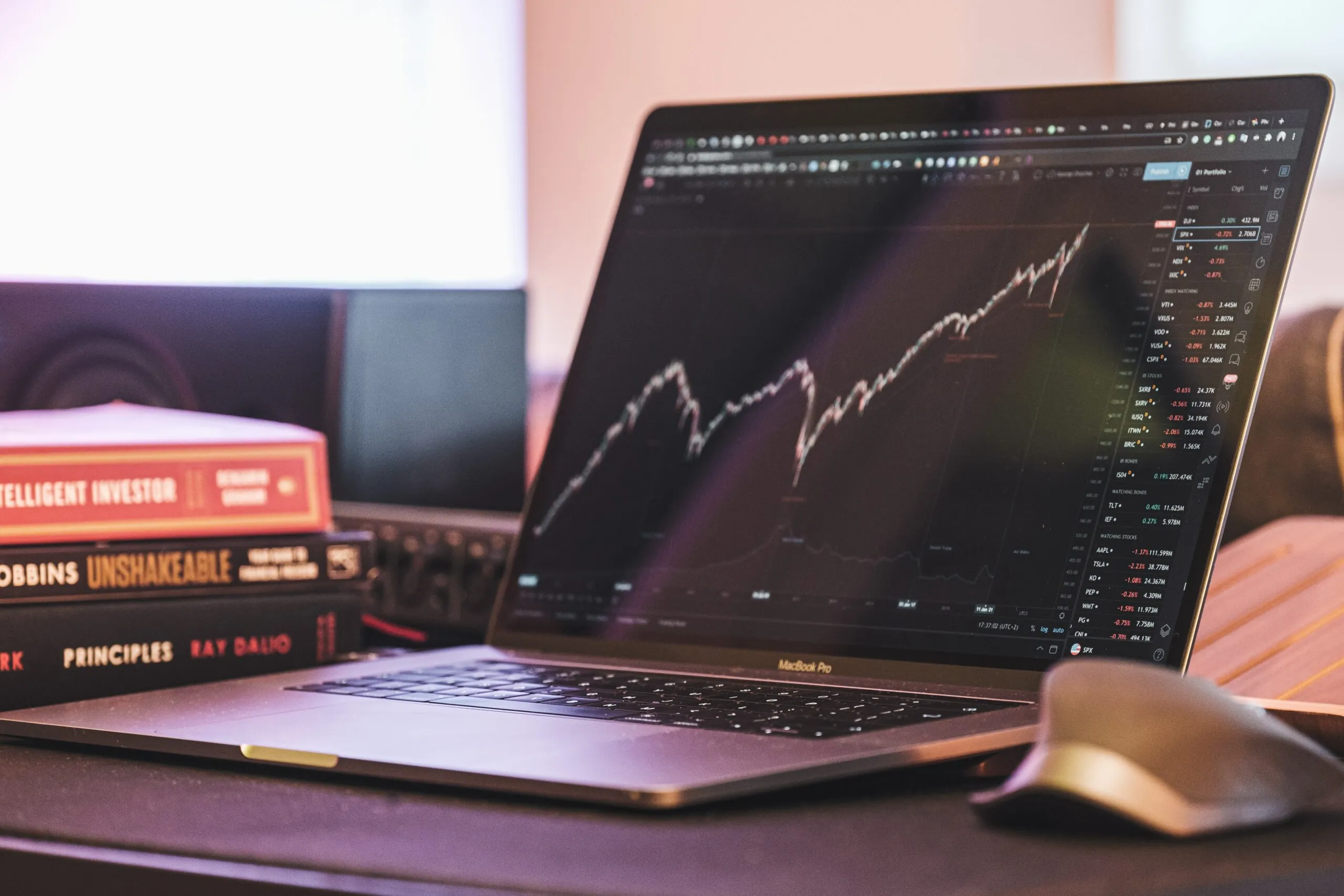 MacBook Pro displaying a stock market chart with financial books stacked nearby on a desk.