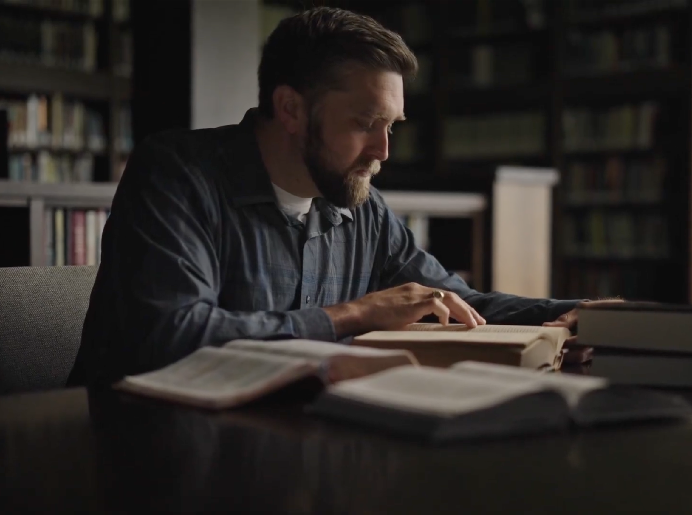 Bearded man reading a book intently at a library table with several open books around him.