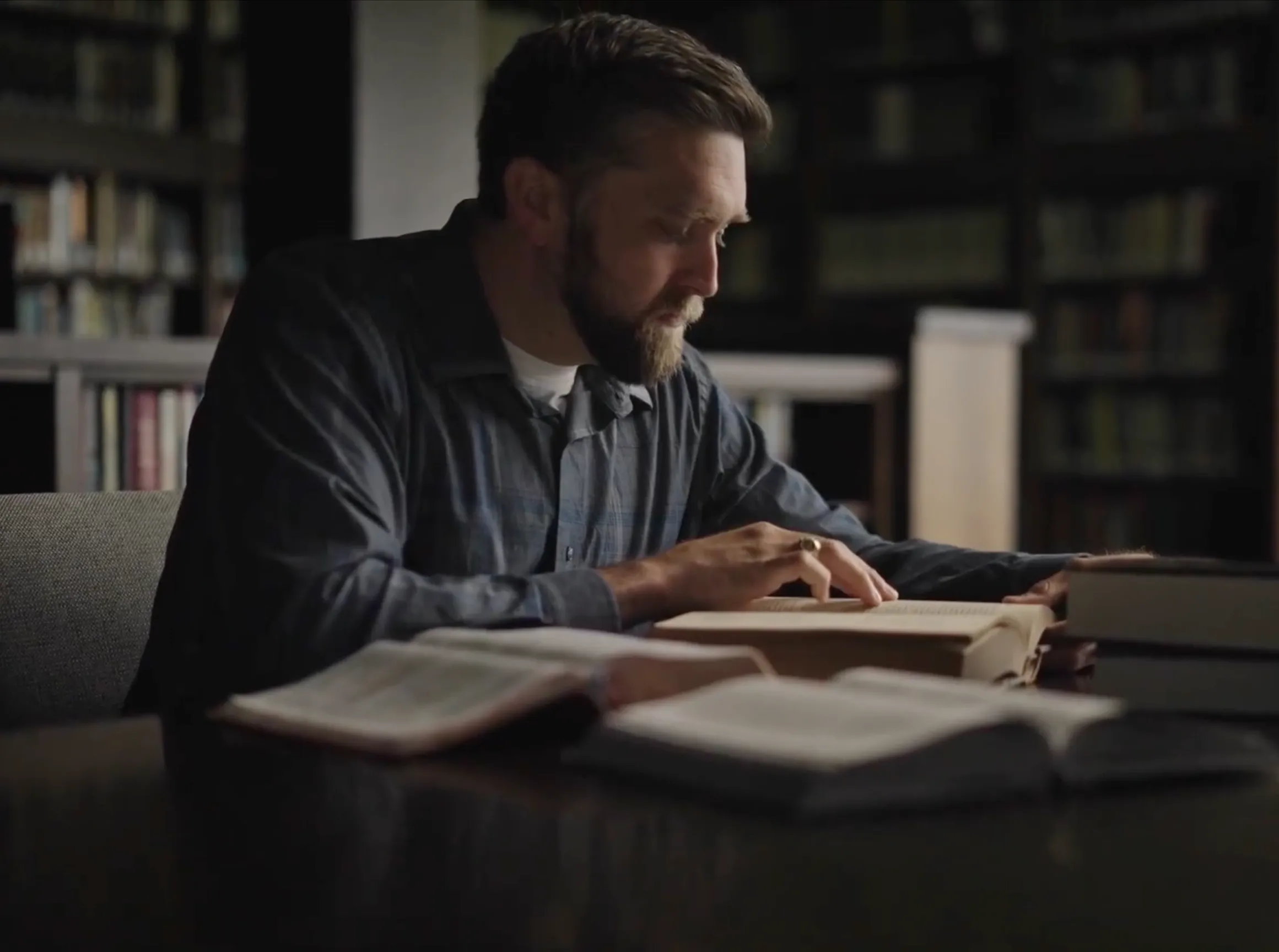 Bearded man reading a book intently at a library table with several open books around him.
