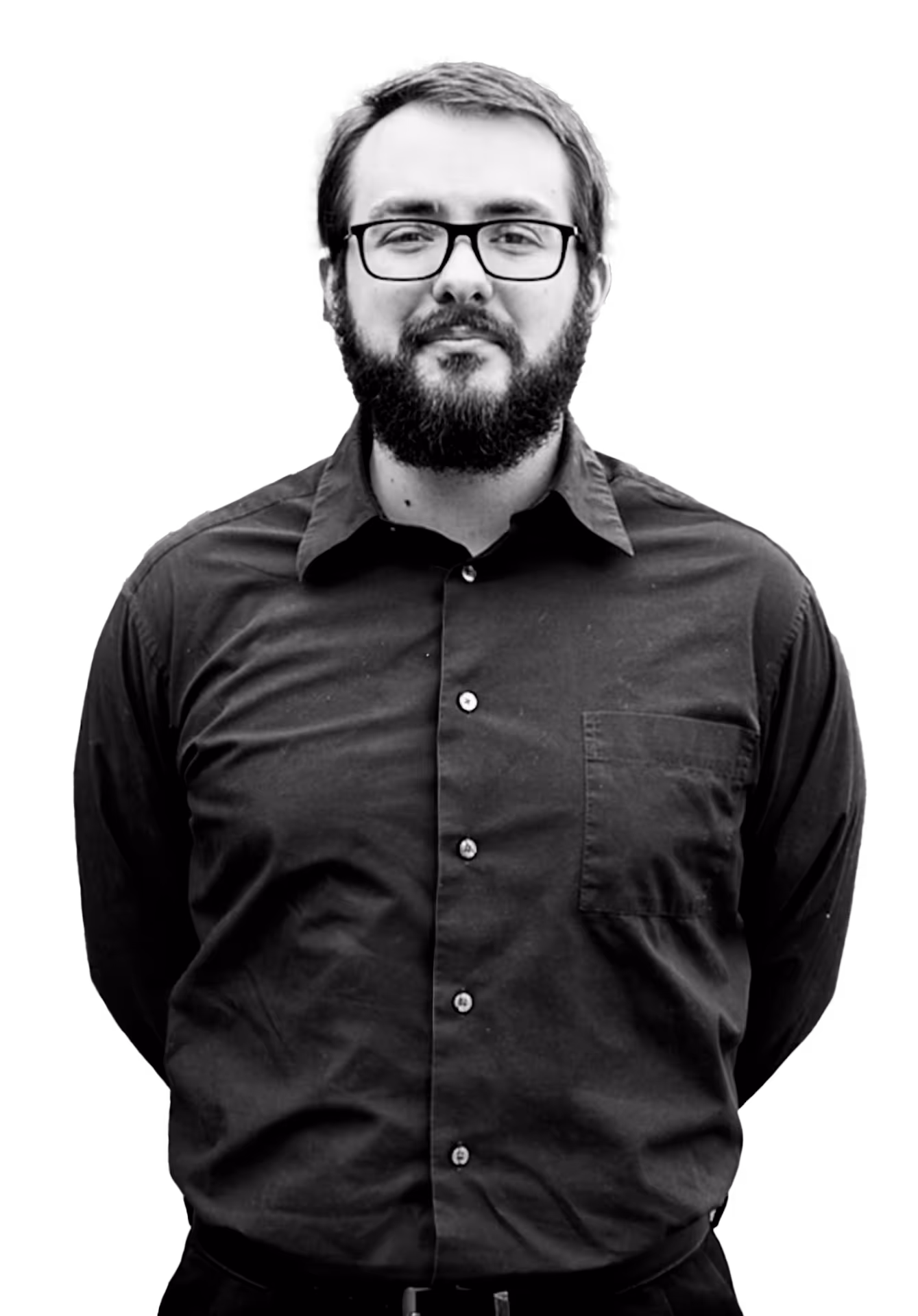 Bearded man with glasses wearing a buttoned black shirt, standing with hands behind his back.