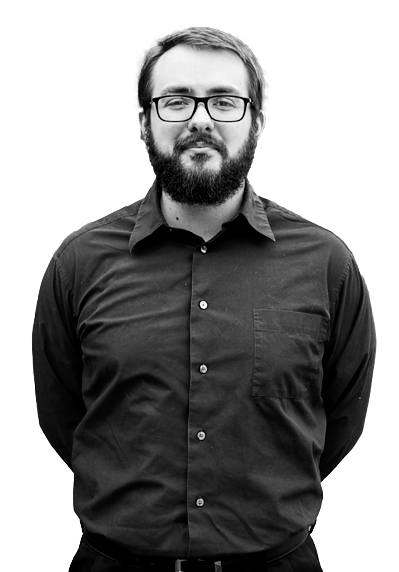 Bearded man with glasses wearing a buttoned black shirt, standing with hands behind his back.