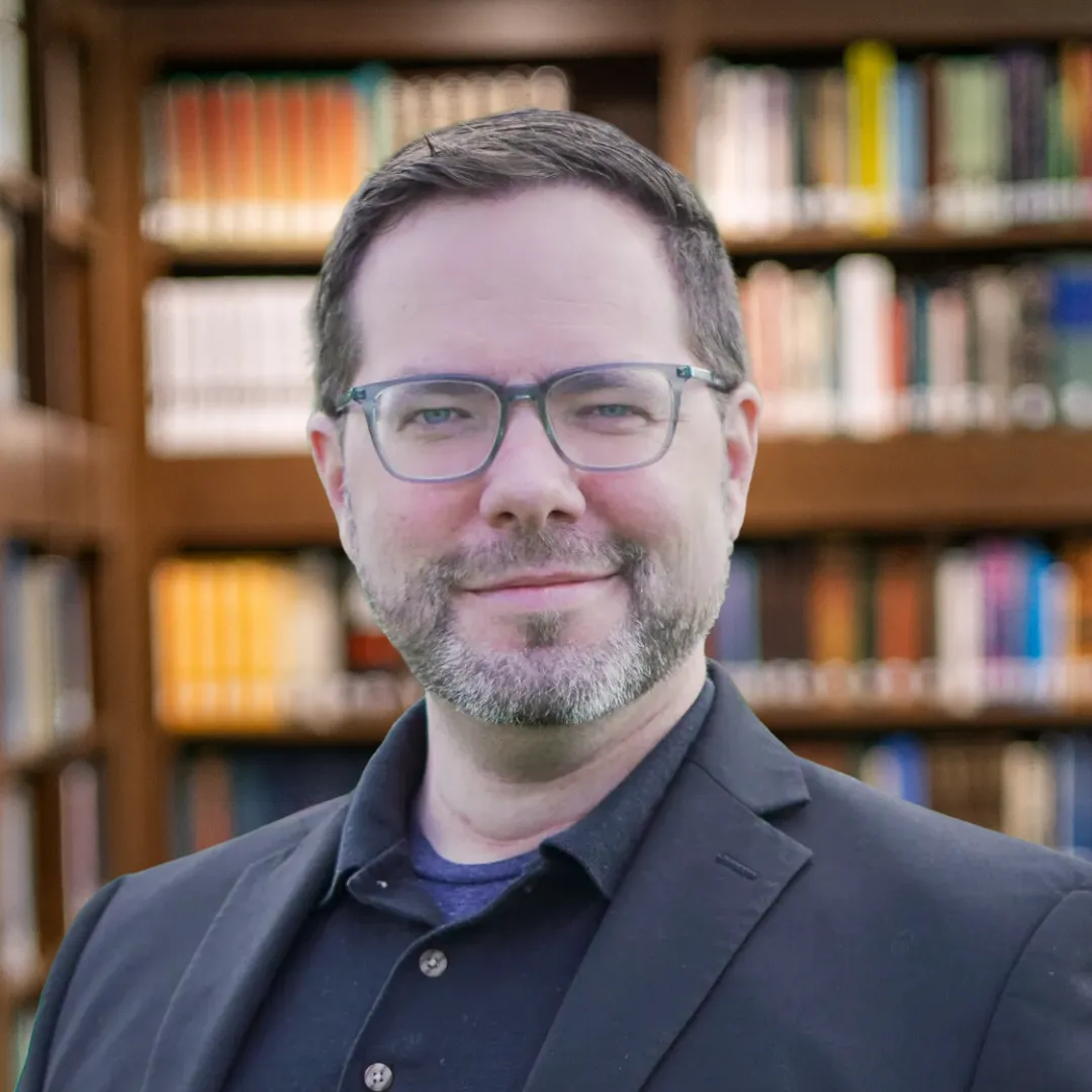 Man with glasses, beard, and short hair wearing a black blazer in front of blurred bookshelves.