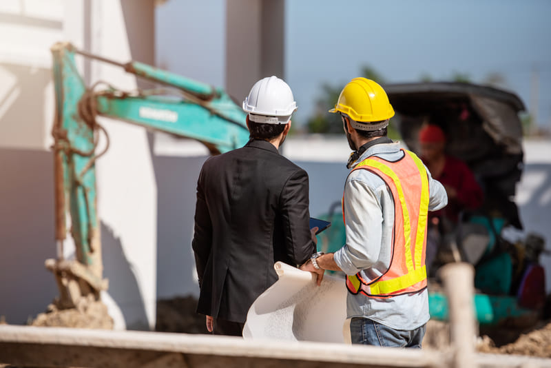 Ingeniero y profesional de obra revisando planos frente a maquinaria de construcción para coordinar actividades, supervisar avances y gestionar la ejecución del proyecto con Diazar.
