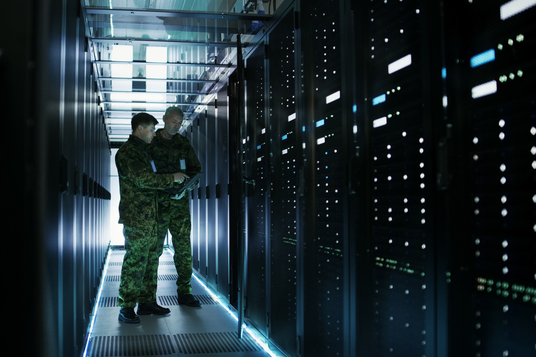 Two military personnel in camouflage uniforms reviewing a tablet inside a dimly lit server room.