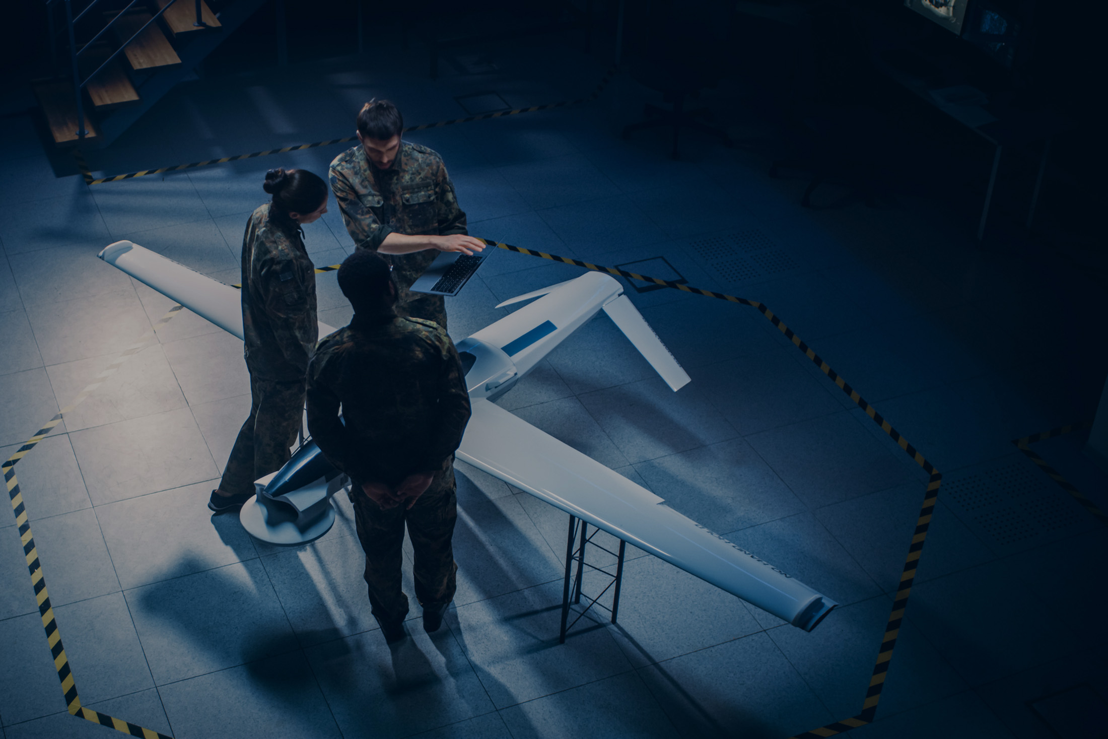 Three military personnel examining a drone model inside a marked area on the floor, with one holding a laptop.