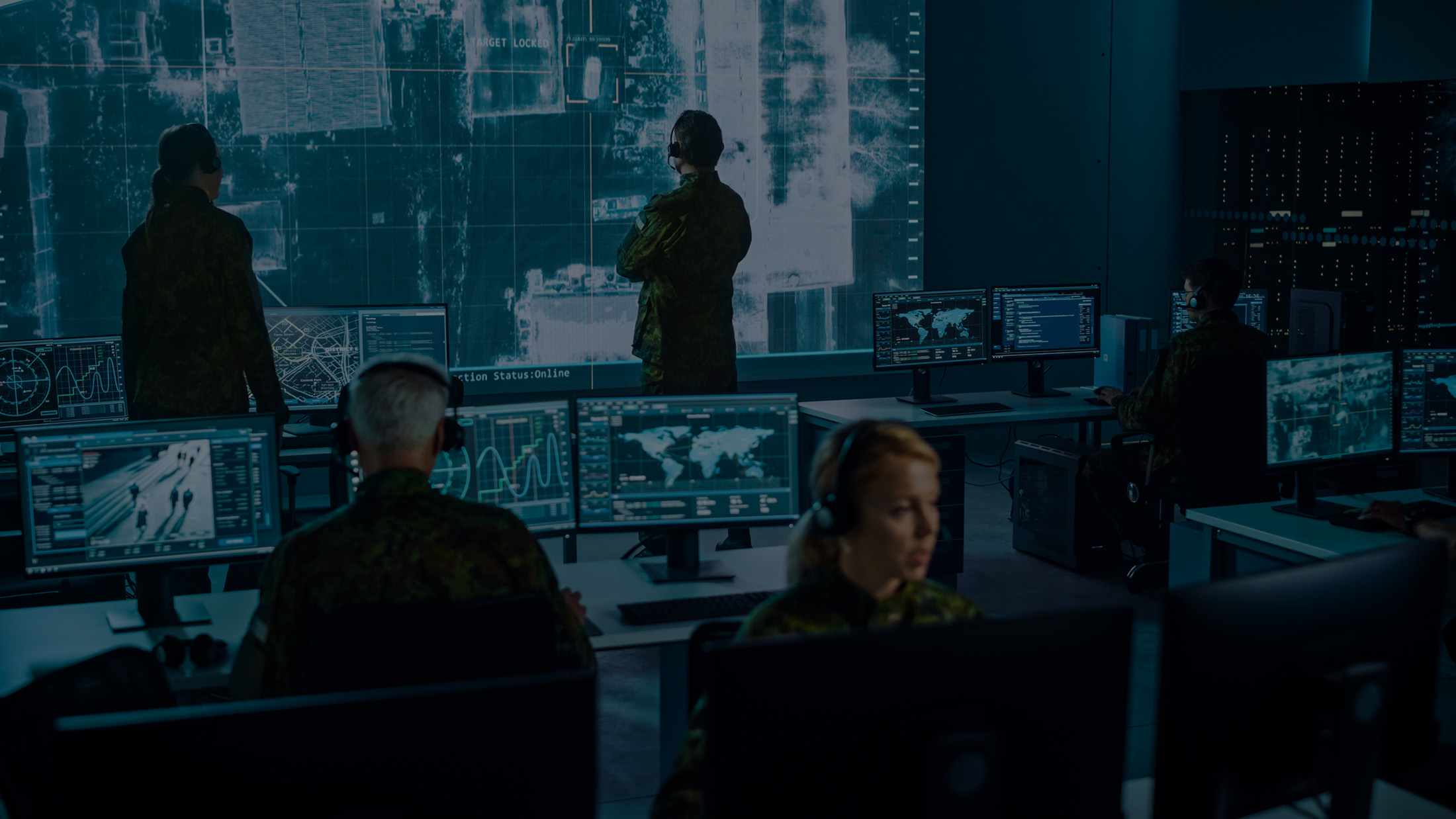 Military personnel in camouflage uniforms monitoring multiple computer screens with maps and data in a command center.